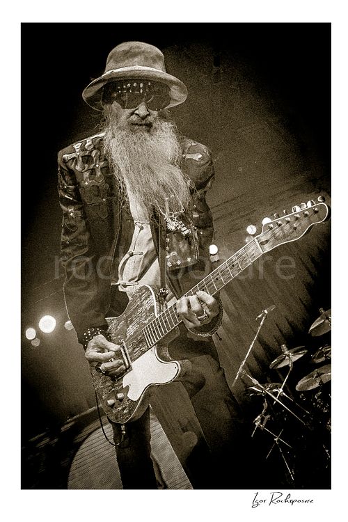 Vertical black and white image of Billy Gibbons of ZZ Top performing live on stage with a Telecaster guitar, wearing a hat, dark glasses, and his signature long beard with dramatic stage light behind him