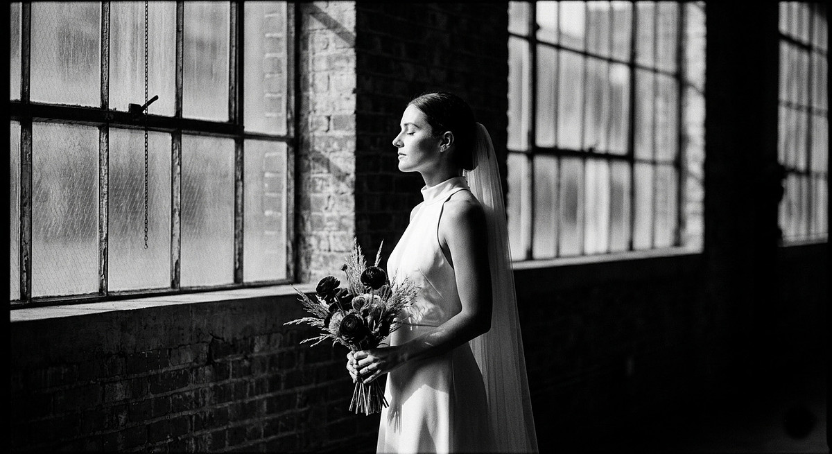 Black and white fine art wedding portrait showcasing a bride in a minimalist gown with dramatic side light (chiaroscuro) against an industrial factory window background.