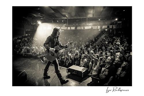 Horizontal black and white image of Tom Keifer performing live on stage with an acoustic guitar, walking toward the crowd under a bright spotlight with fans reaching forward