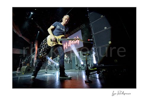 Horizontal color image of Phil Collen of Def Leppard performing live on stage in a dramatic low-angle shot, playing guitar with the Def Leppard logo visible in the background