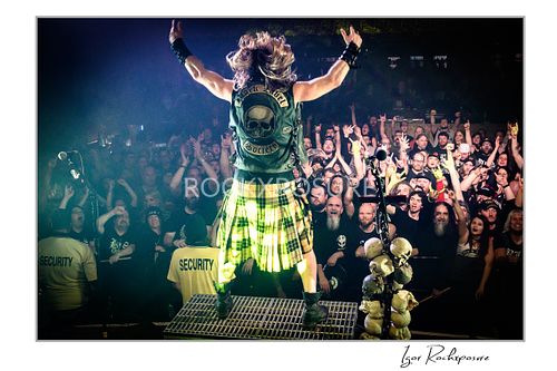 Wide shot from behind Zakk Wylde on stage with arms raised, wearing a kilt and vest, cheering the large crowd.