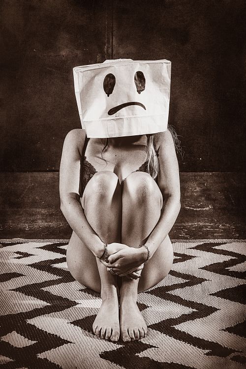 Femme assise au sol en lingerie avec un sac en papier sur la tête affichant un visage triste, illustrant l’épuisement émotionnel et la solitude intérieure malgré une façade sociale