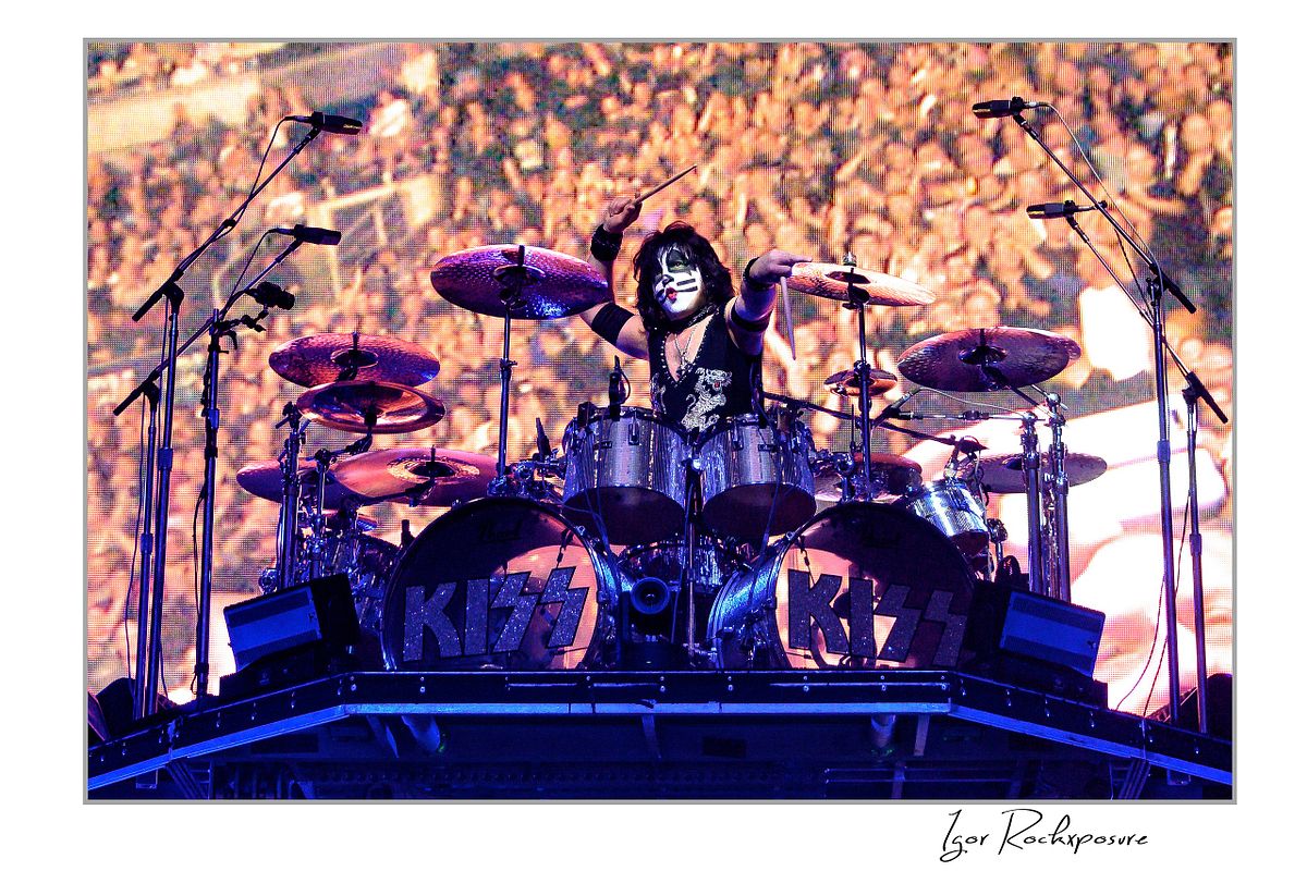 Eric Singer of KISS playing the drum kit on a raised stage platform with a massive crowd image displayed on the arena screen behind him under warm concert lighting