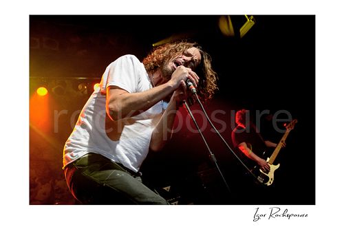 Horizontal color image of Chris Cornell and Ben Shepherd of Soundgarden performing live on stage, with Cornell at left singing into a microphone and Shepherd at right playing bass under warm concert lighting