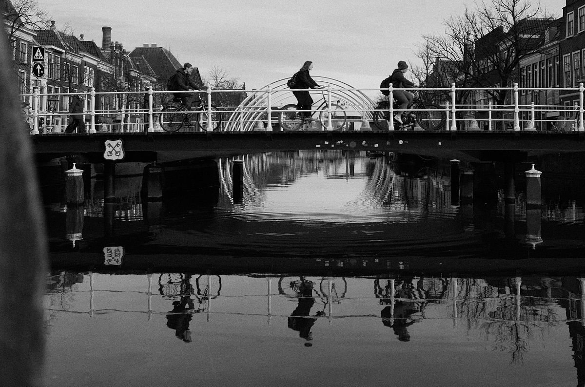 Cyclists crossing a bridge over a canal in the Netherlands with their reflections in the water, captured by photographer Sandeep Gajula