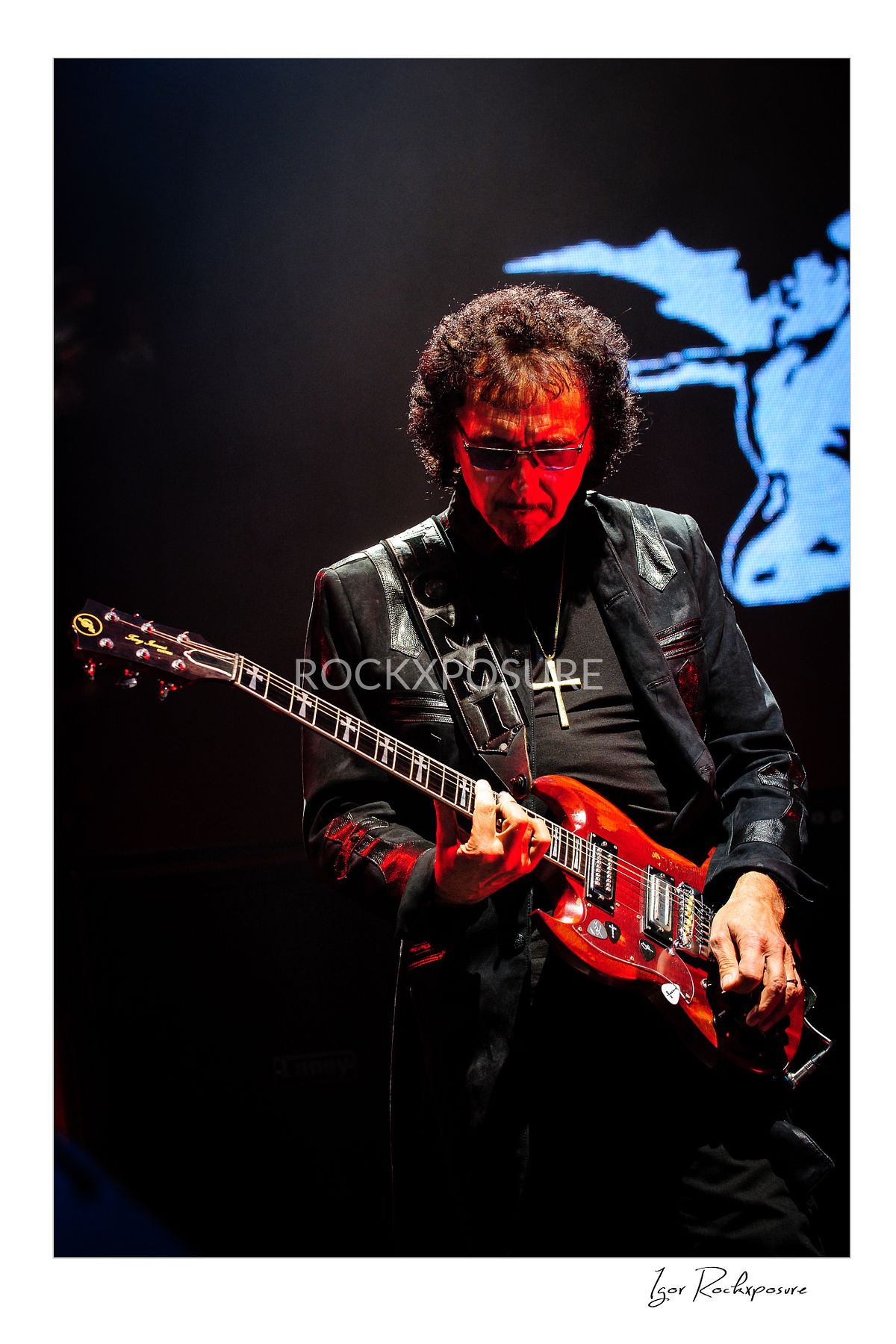 Tony Iommi of Black Sabbath playing electric guitar on stage under dramatic red concert lighting with dark stage backdrop