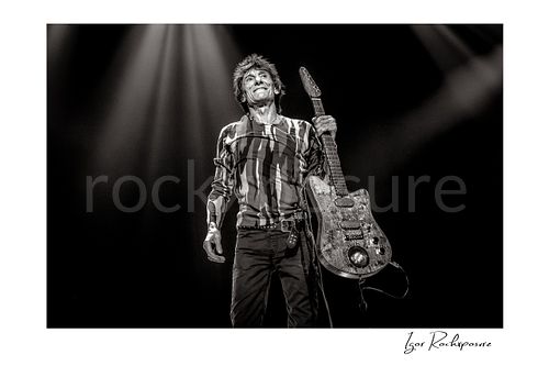 Horizontal black and white image of Ronnie Wood of The Rolling Stones holding an electric guitar beneath dramatic stage spotlights