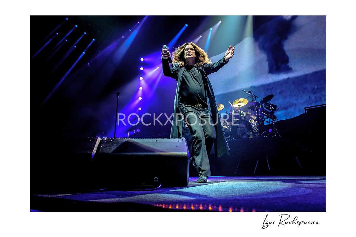 Ozzy Osbourne of Black Sabbath stepping forward across the stage with one arm raised under purple and blue concert lighting during a live performance