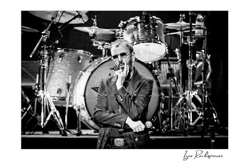 Horizontal black and white image of Ringo Starr standing onstage before the drum kit in sunglasses with one finger raised to his lips