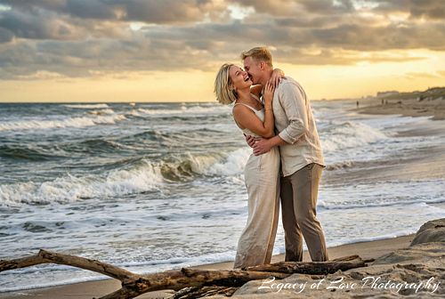 A surprised woman and a man in a passionate embrace on a St. Augustine, Florida beach at sunset after a successful marriage proposal. The couple is standing near a large piece of driftwood.