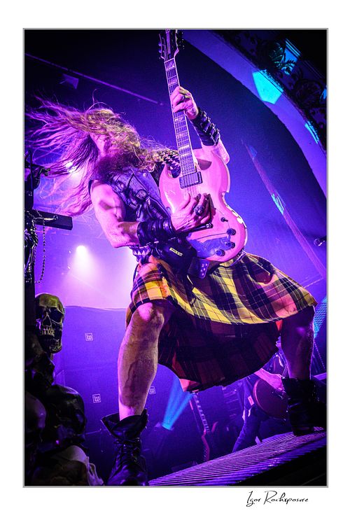 Low-angle vertical shot of Zakk Wylde standing with legs wide apart on a stage riser, wearing a kilt and playing guitar with his hair flying under vibrant purple and blue stage lights.