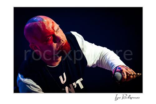 Horizontal color image of Ivan Moody of Five Finger Death Punch in a tight onstage close-up with microphone in hand under red and blue concert light