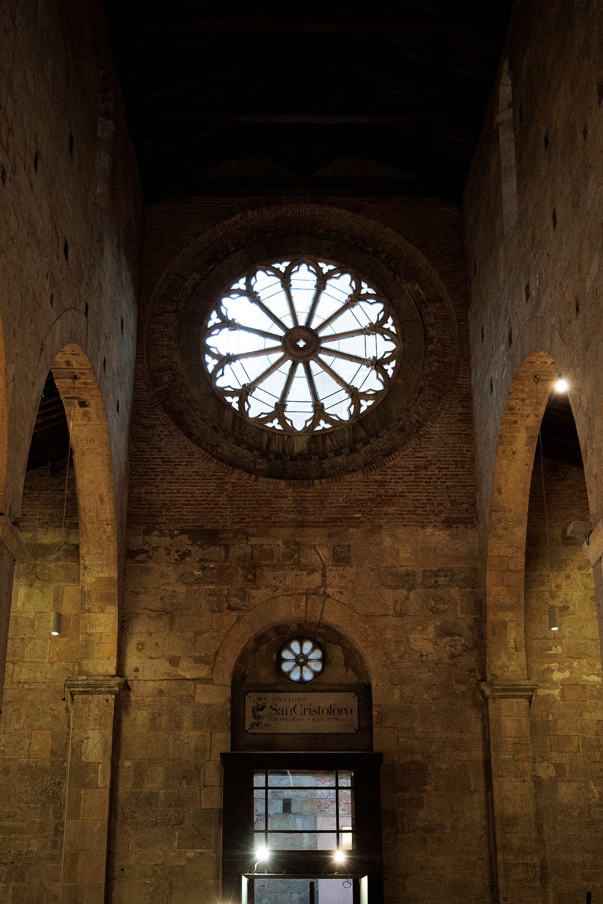 Interior view of Museo San Cristoforo in Lucca, Italy, showing a high stone wall with a large gothic circular window at the top, a smaller round floral window below and a doorway framed by a sign reading &ldquo;Museo San Cristoforo&rdquo;, with soft natural and artificial light revealing the rough, aged stone texture and creating a quiet, atmospheric mood.