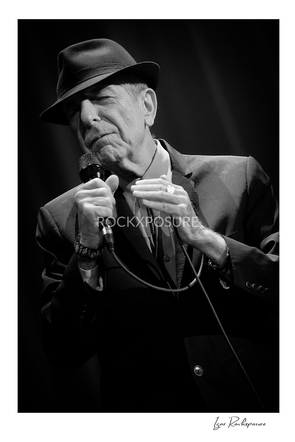 Vertical concert photography of Leonard Cohen singing into a microphone in black and white photography under stage lighting