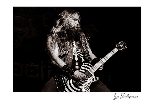 Black and white shot of Zakk Wylde wearing a denim vest, playing his signature bullseye guitar on stage against a dark background.
