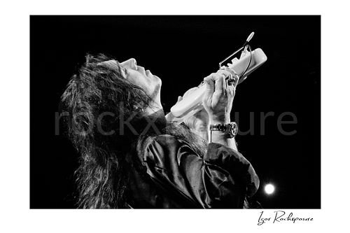Horizontal black and white image of Yngwie Malmsteen in a dramatic live performance close-up, head tilted back while holding a white guitar against his neck under dark stage light