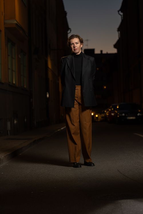 Kvinna i svart kavaj står på mörk stadsgata i skymningsljus, redaktionellt porträtt