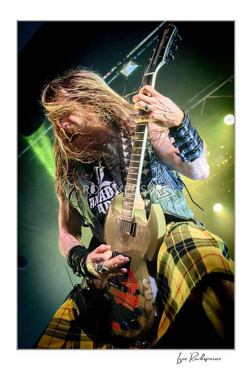 Vertical shot of Zakk Wylde playing guitar on stage, wearing a yellow plaid kilt and denim vest, with his long hair falling forward covering his face.