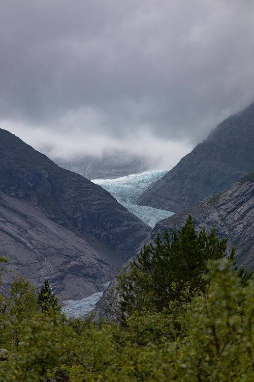 Glacier bleu encaissé entre deux montagnes rocheuses en Norvège, avec brume et ciel couvert en été.