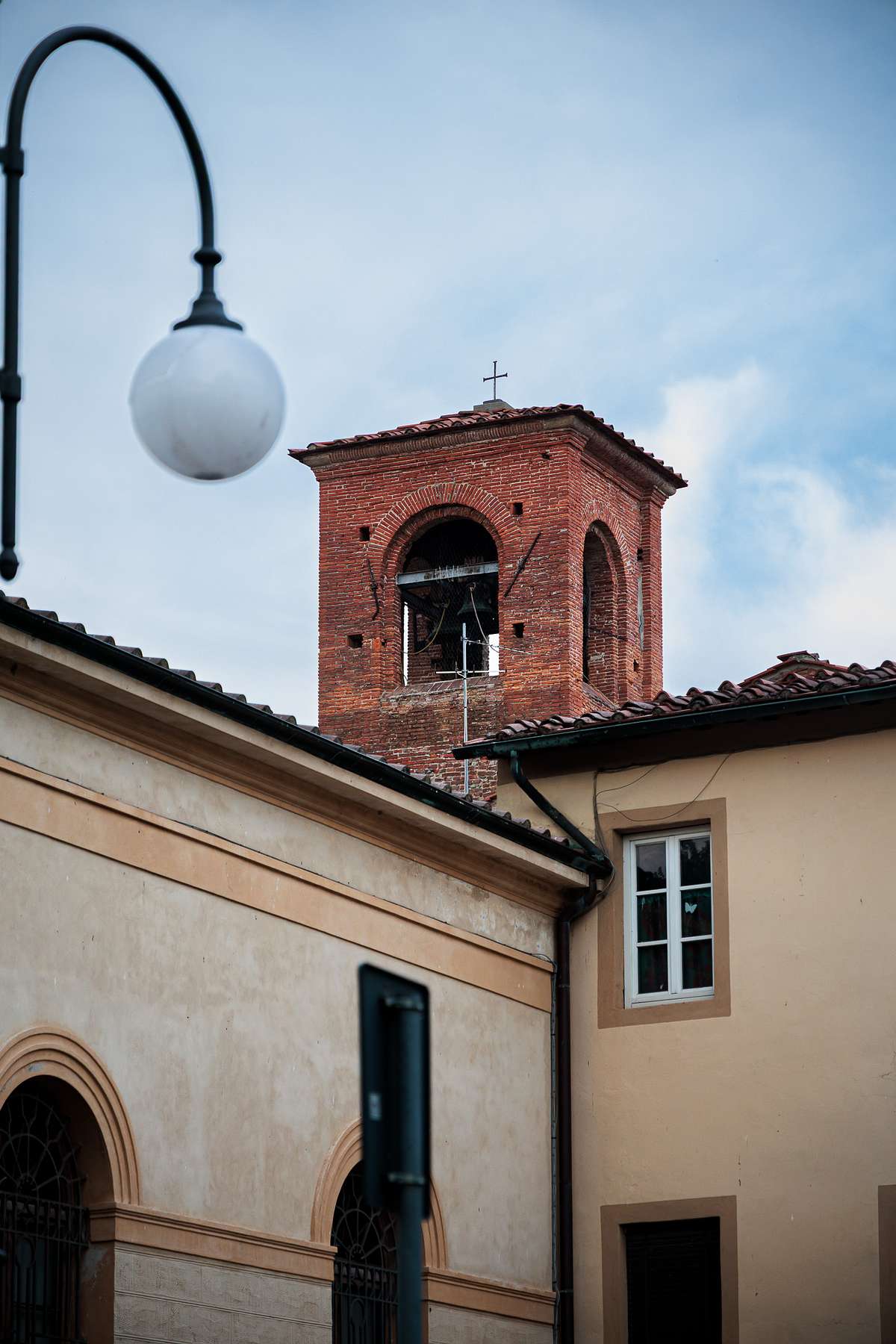 Street view of a red brick bell tower with a cross on top rising above light-colored buildings in Lucca, Italy, with a street lamp, sloped roofs and a partly cloudy sky creating a calm urban scene.