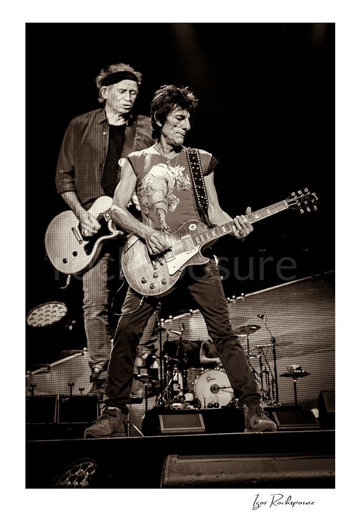Vertical black and white image of Ronnie Wood and Keith Richards of The Rolling Stones performing on stage with guitar and giant backdrop screen