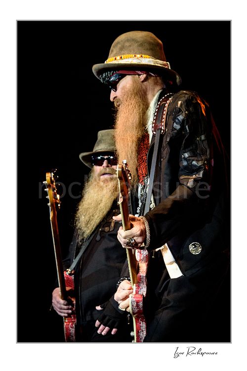 Vertical color image of Dusty Hill and Billy Gibbons of ZZ Top performing live on stage with red guitars, wearing signature hats, dark glasses, and long beards against a dark background