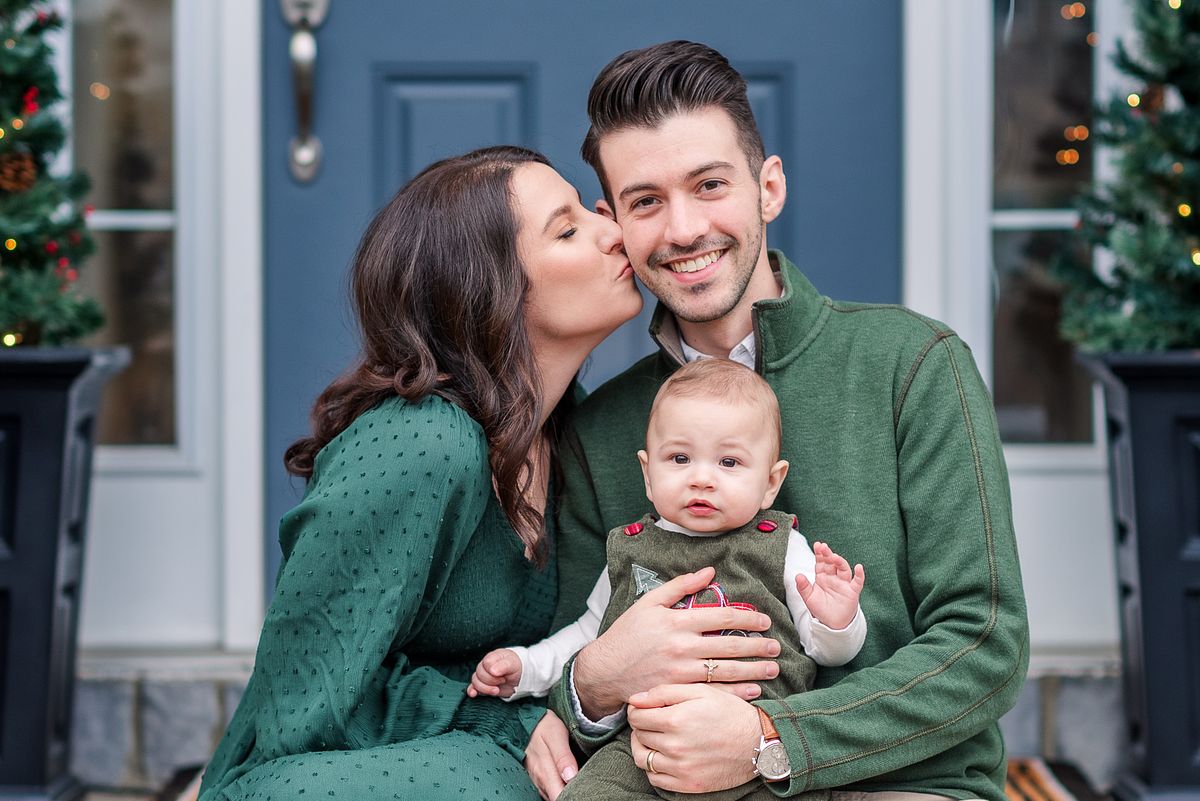Wife kissing her husband on the cheek while he holds their baby and smiles at Christmas time with Cranberry Township, PA newborn photographer