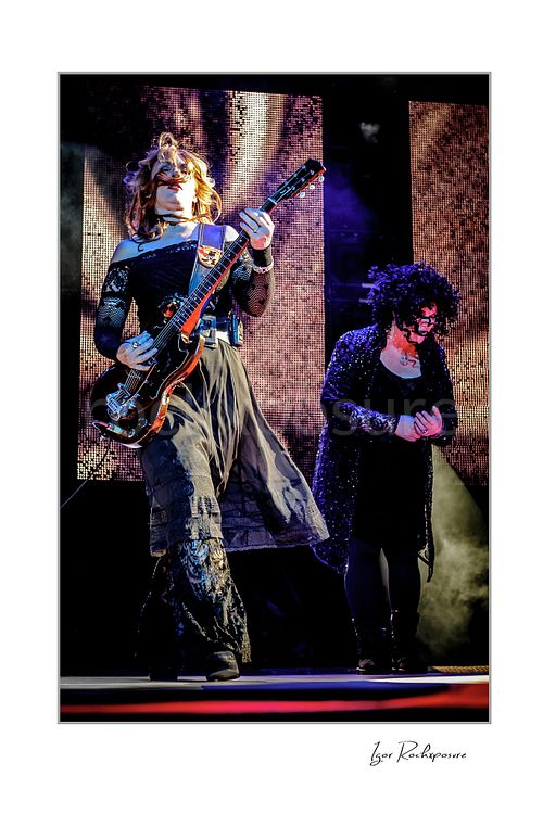 Vertical color image of Nancy Wilson and Ann Wilson performing live on stage during a Heart concert with electric guitar, dark stagewear and bright concert screens