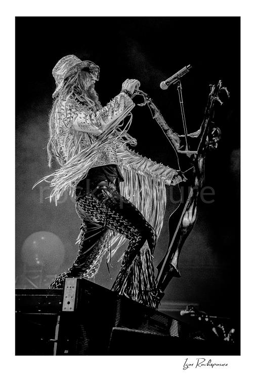Vertical black and white image of Rob Zombie performing live on stage in a fringed jacket and hat with a microphone stand, flowing dreadlocks, and dramatic side profile