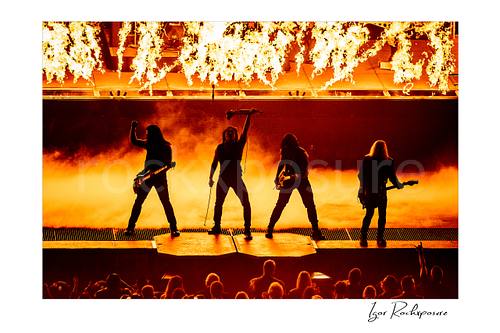 Horizontal color image of Trans Siberian Orchestra performing in silhouette with guitars, raised microphone stand, flames, and stage smoke