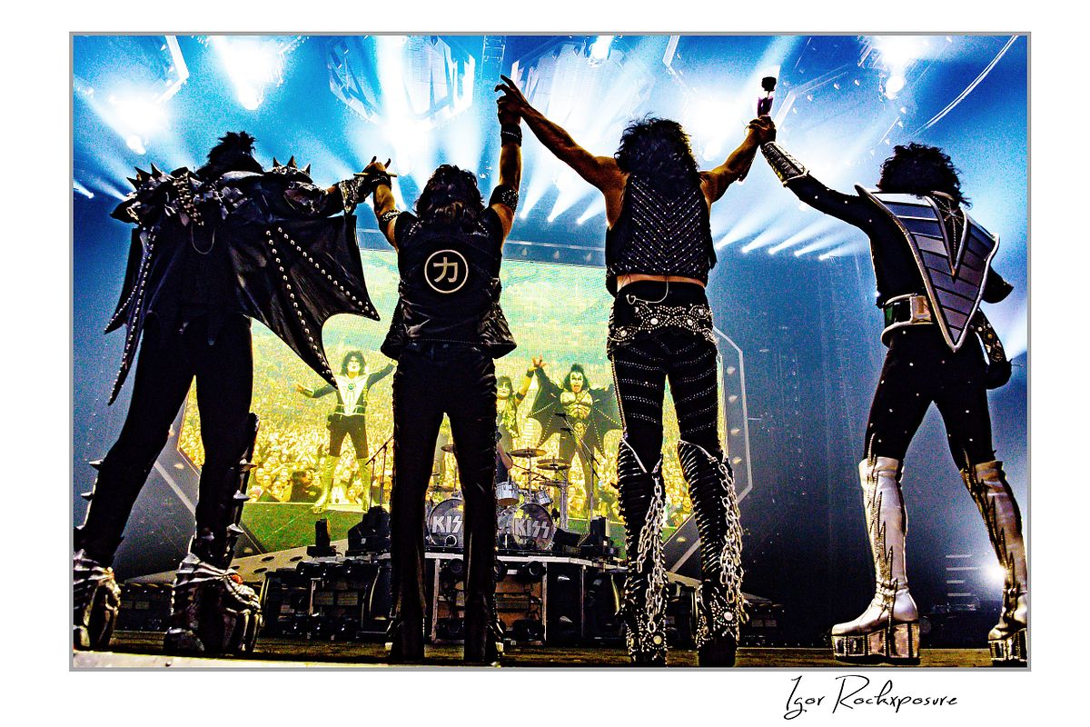 Horizontal color photograph of the KISS band standing onstage with their backs to the camera holding hands in the air while facing a massive stage screen that displays their own image, creating a dramatic mirrored moment in front of the crowd
