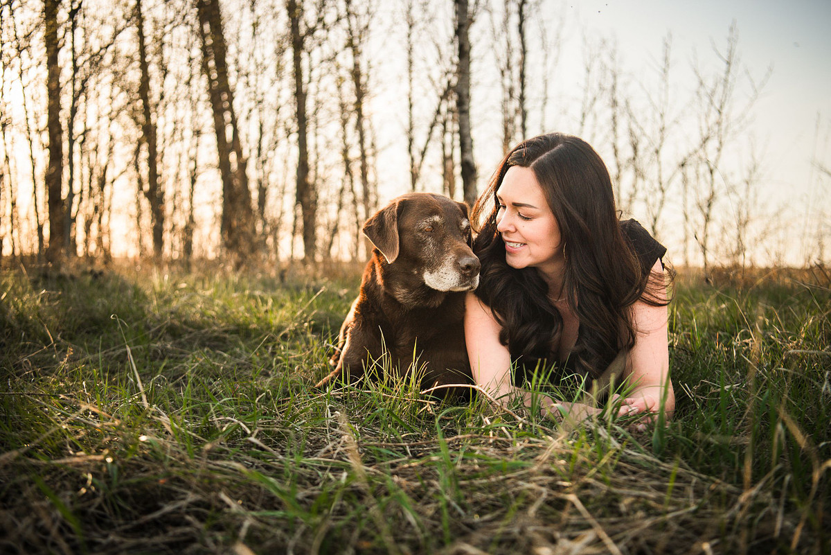 a chocolate lab and a brunette look adoringly into each other's eyes with grass in the foreground and trees in the back. the pair are positioned closley.