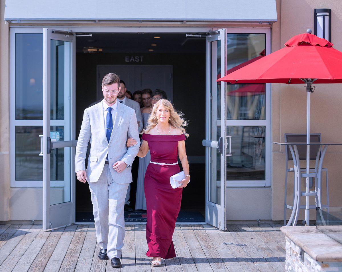 the groom and his mom walking out on the terrace at the hyatt, dewey beach. the sun is shining bright, the mom is wearing a red of the shoulder dress the groom is wearing a grey suite