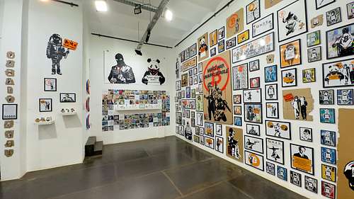 Panoramic view of the Street Cyber "Art of Revolution" exhibition in a gallery, showcasing a wall densely covered with framed stencil artworks from various collections, including "Banditos," "Bitcoin Squad," "Bonanza," and iconic pieces like "Make War Unaffordable" and "Born Debt-Free." The artworks blend pop culture and street art to convey powerful messages about Bitcoin, financial freedom, and social change. This image captures the immersive scale and diverse themes of the exhibition.
