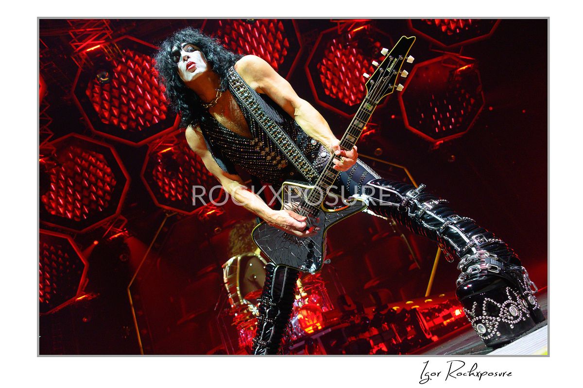 Paul Stanley of KISS performing a wide power stance while playing guitar under intense red stage lighting with dramatic circular stage visuals
