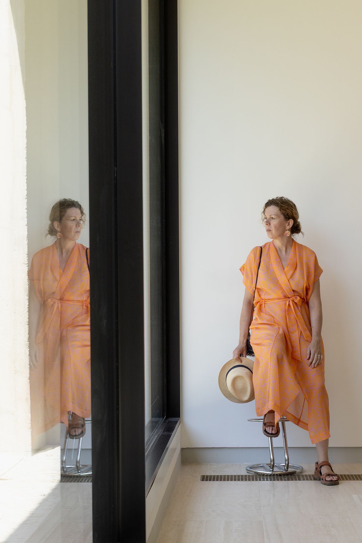 Kvinna i orange kl&auml;nning sitter p&aring; en pall och h&aring;ller en hatt; hennes spegling syns i ett f&ouml;nster.