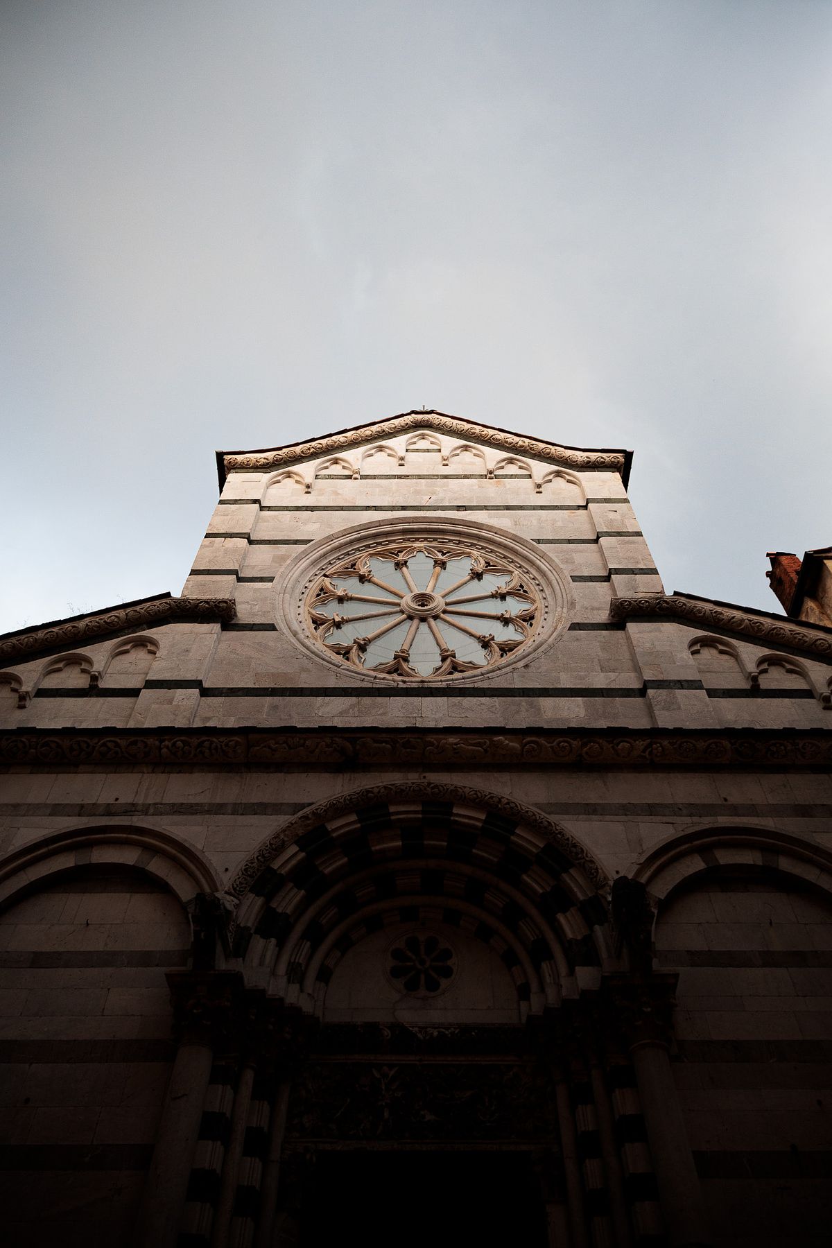 Low-angle photo of a historic Tuscan church facade with light stone stripes, an ornate circular window and a peaked roof, shot under an overcast sky creating a moody atmosphere.