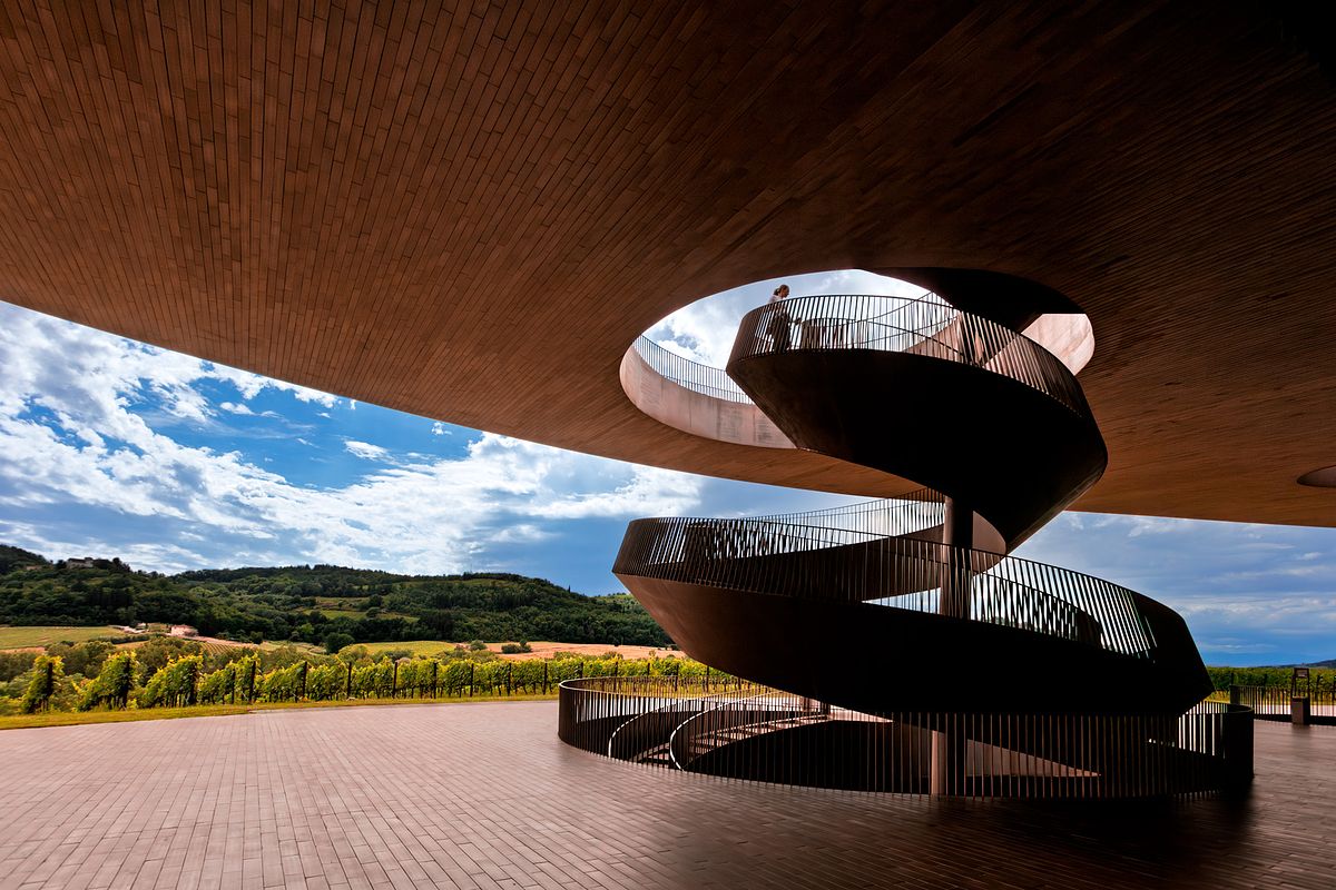 A dramatic low-angle view of a weathered Cor-Ten steel spiral staircase winding through a circular opening in a terracotta ceiling toward a blue sky at the Antinori winery in Tuscany.