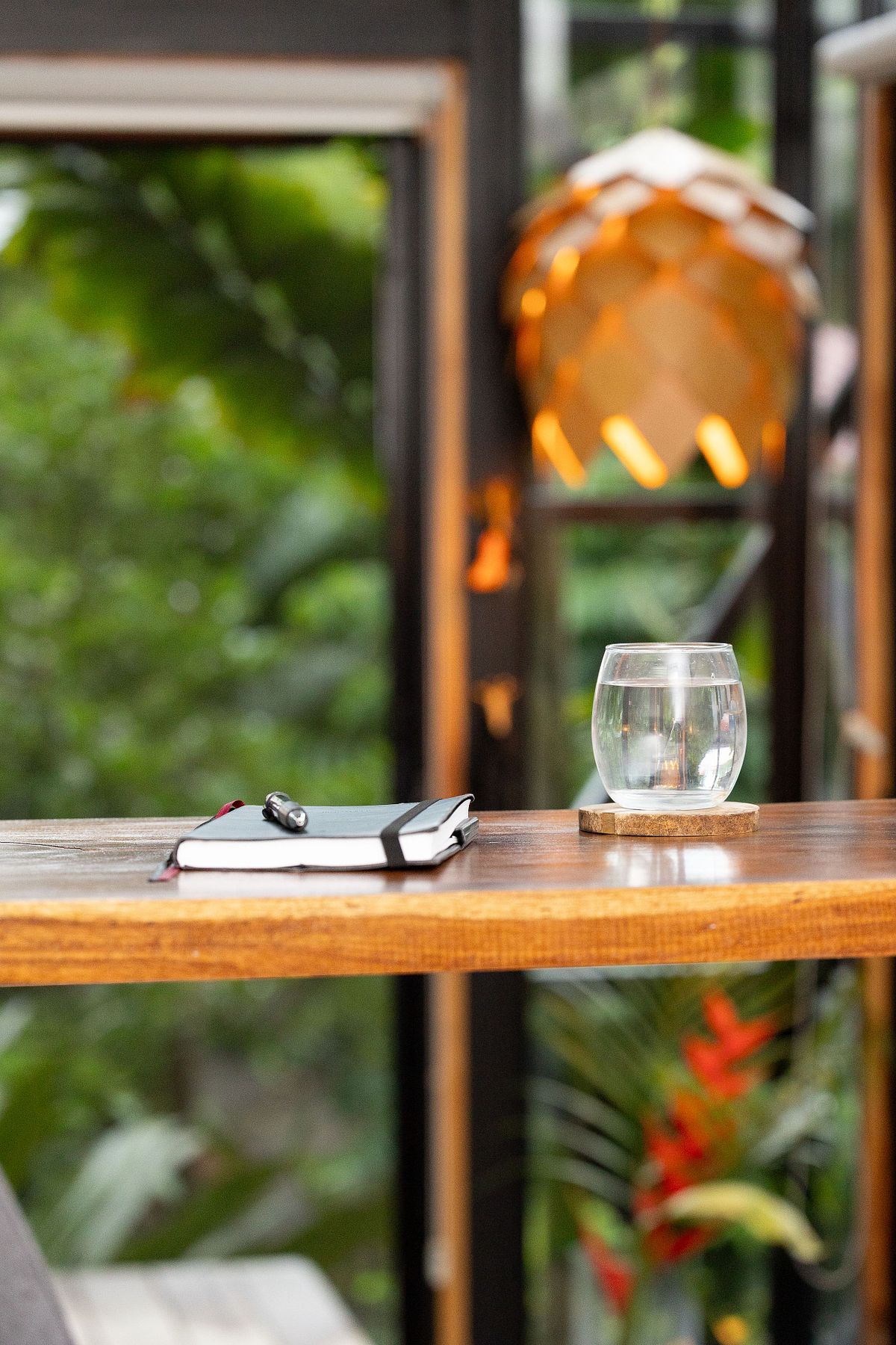 Redaktionell st&auml;mningsbild med anteckningsbok, penna och vattenglas p&aring; tr&auml;bord i tropisk gr&ouml;nska.