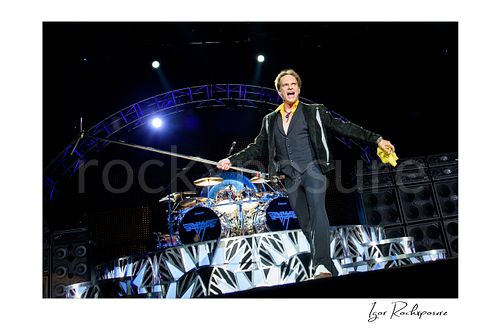 Horizontal color image of David Lee Roth of Van Halen performing on stage in front of Alex Van Halen’s drum kit and speaker stacks