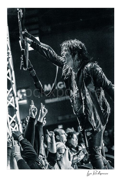 Vertical black and white image of Tom Keifer performing live at the edge of the stage, raising his black guitar overhead toward the crowd in a fierce concert moment