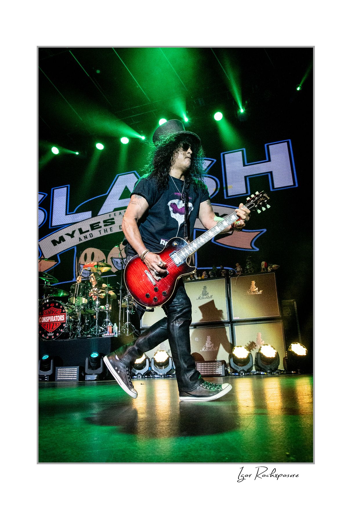 Vertical concert photography of Slash playing electric guitar in a wide stance under green stage lighting with Slash stage backdrop