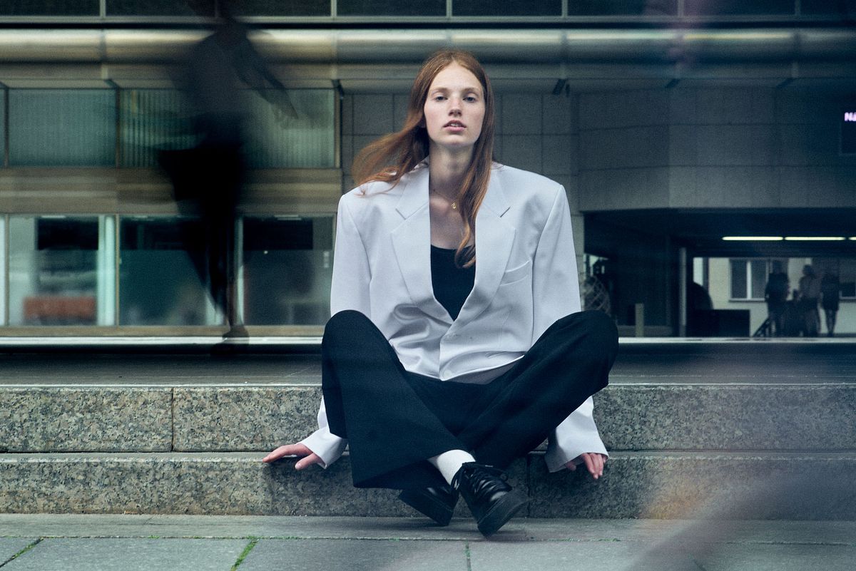 Contemporary fashion portrait of a woman in an oversized white blazer sitting in an urban environment with motion blur, photographed by Gabriel Matula.