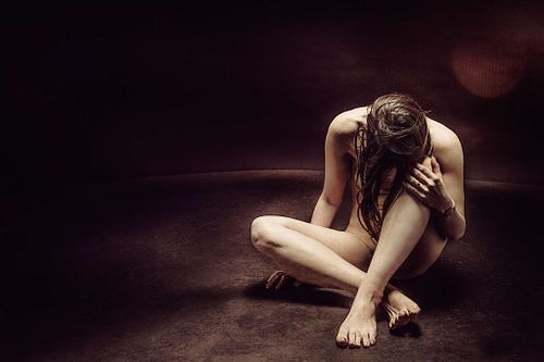 Femme nue assise au sol dans un décor sombre, penchée vers l’avant, symbolisant la culpabilité et les conséquences du karma