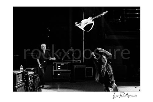 Horizontal black and white image of Yngwie Malmsteen tossing his white guitar high across the stage toward legendary guitar tech Thomas Nordegg, captured in a dramatic live performance moment