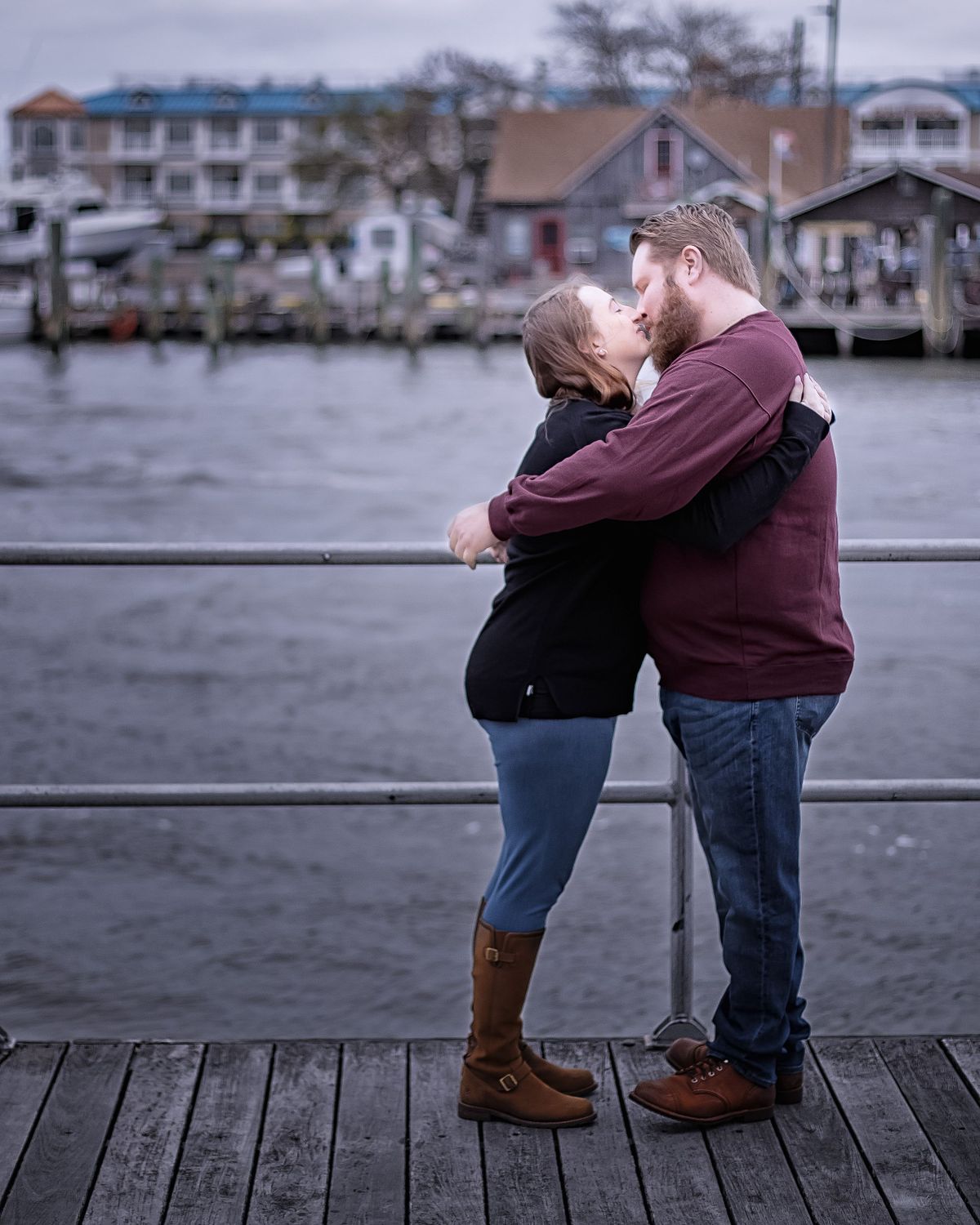 couple embracing on the boardwalk in lewes, delaware, both are wearing jeans and she is wearing a black sweater and he is wearing a tan sweater. they are kissing, the canal is in the background with houses on the other side
