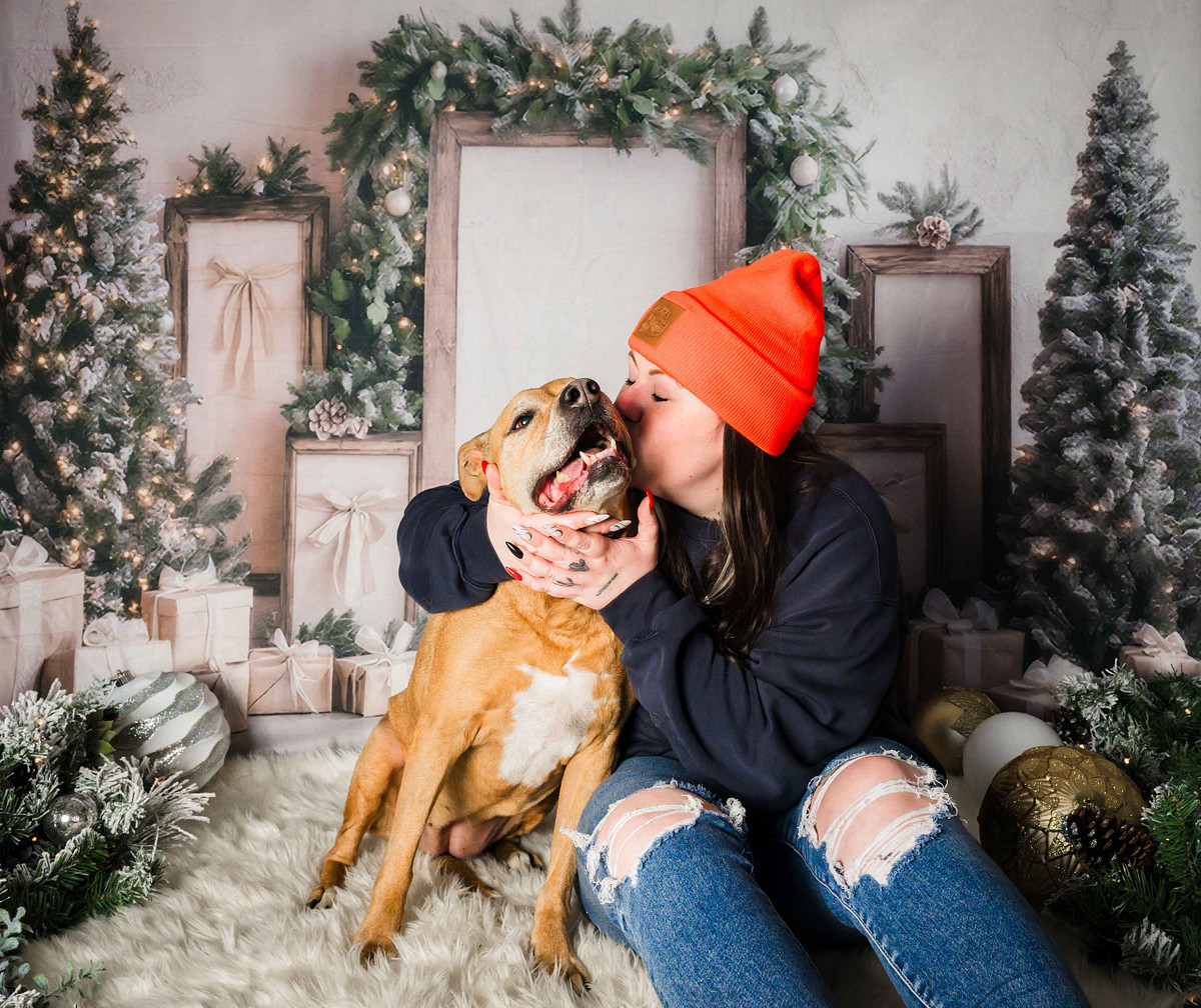woman in an orange beanie kissing her senior tan rescue dog during a cozy Christmas themed pet photo session, surrounded by lit holiday trees, wrapped presents, and frosted greenery, in Calgary at Sleep Rover