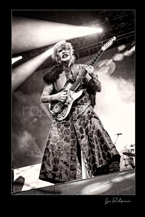 Vertical black and white image of John 5 performing live on stage with an electric guitar, theatrical makeup, patterned stage outfit, and bright backlighting