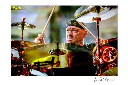Horizontal color image of Neil Peart performing at his drum kit during a Rush concert, framed by cymbals with raised drumsticks and intense expression