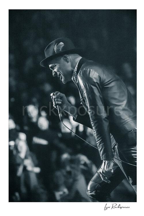 Vertical black and white image of Gord Downie of The Tragically Hip leaning forward with microphone in hand during a live performance, wearing a hat and leather jacket with the crowd softly blurred behind him
